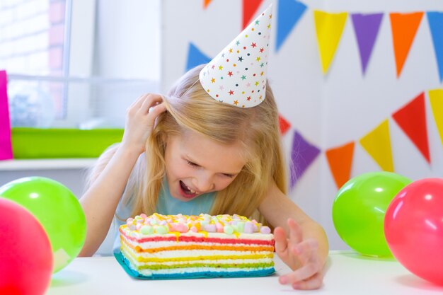 Menina loira caucasiana espiando por trás de um bolo de aniversário com uma cara engraçada na festa de aniversário