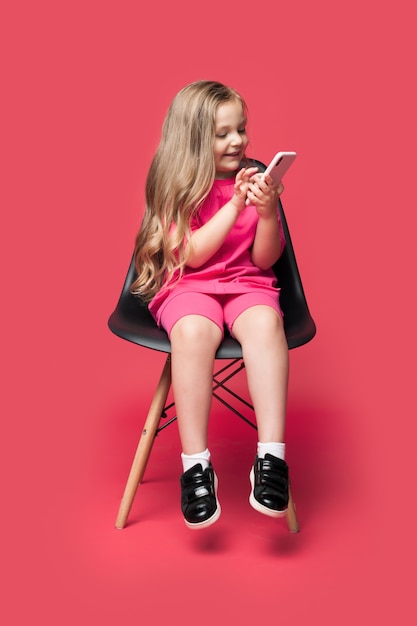 Menina loira caucasiana está sentada em uma cadeira sorrindo usando um telefone em uma parede vermelha do estúdio