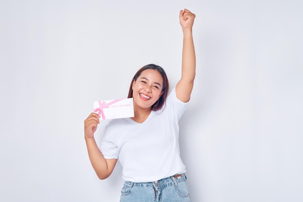 Menina loira chocada, asiática, vestindo camiseta branca casual, segurando vale-presente, comprovante de cupom, levantando o punho, comemorando boa sorte, isolado no fundo branco Conceito de estilo de vida de pessoas