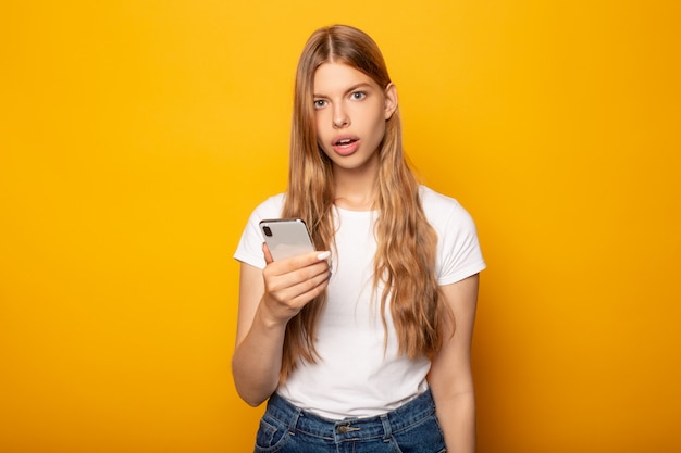 menina loira chocada usando smartphone isolado em amarelo