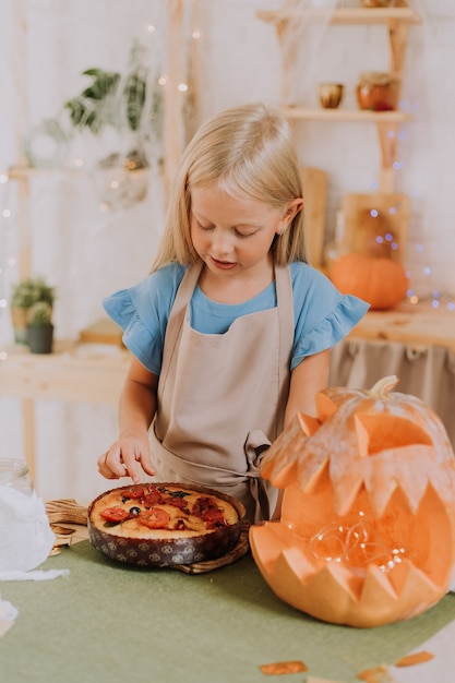 menina loira com avental na cozinha decorada com abóboras para o Halloween preparando uma torta de focaccia