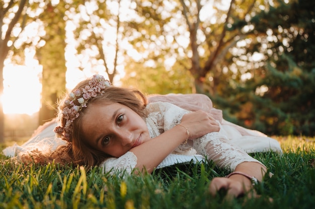 Foto menina loira com cabelo encaracolado com vestido de comunhão deitada na grama para fotos