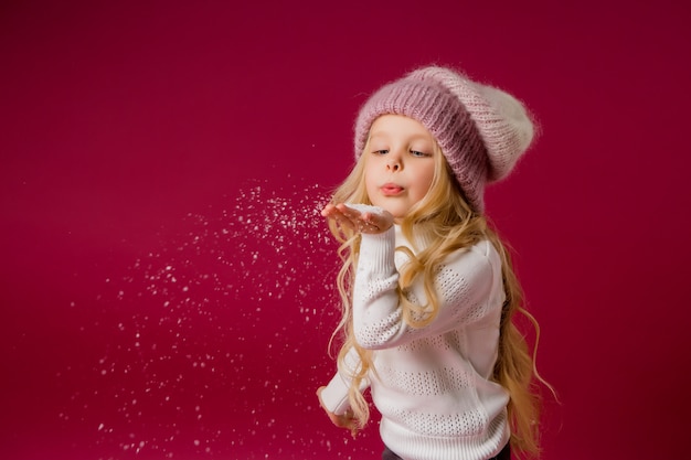 Menina loira com chapéu de malha e blusa brinca com a neve