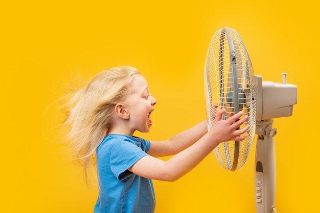 Menina loira com movimento no cabelo do vento segurando ventilador de suporte e gritando Criança e circulador de ar no fundo amarelo