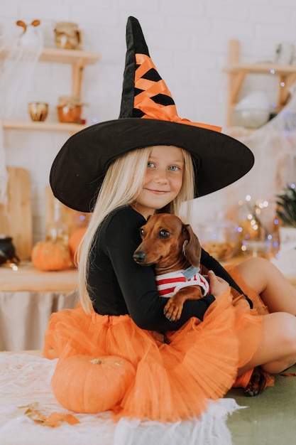 menina loira com uma fantasia de bruxa saia fofa laranja segurando um dachshund anão no colo Halloween