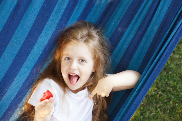 Menina loira de cabelo comprido come um morango no jardim deitado em uma rede azul
