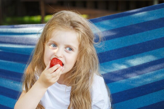 Menina loira de cabelo comprido come um morango no jardim sentado em uma rede azul