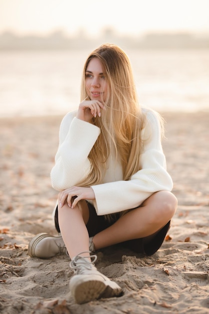 Menina loira de cabelos compridos sentada na praia de areia na manhã de outono