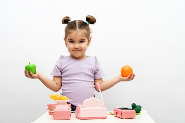 Menina loira de camiseta roxa segura frutas na mão joga na loja infantil brinquedo caixa registradora escala legumes e frutas na cesta na comida saudável de fundo branco