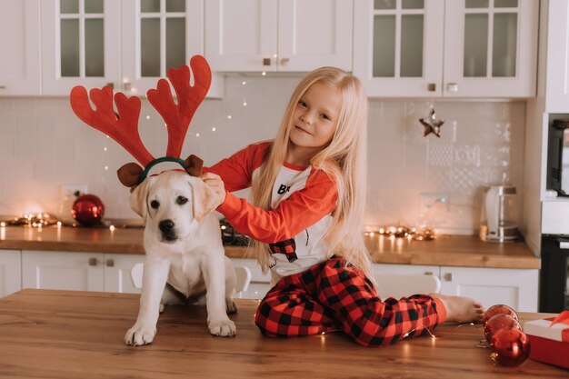 Menina loira de pijama vermelho de Natal coloca chifres de veado de brinquedo na cabeça de um cachorro. interior festivo. criança brinca com seu amado animal de estimação. estilo de vida. espaço para texto. Foto de alta qualidade