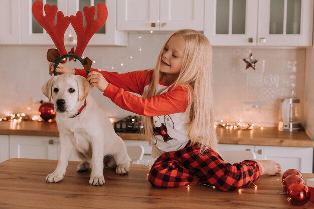 Menina loira de pijama vermelho de Natal coloca chifres de veado de brinquedo na cabeça de um cachorro. interior festivo. criança brinca com seu amado animal de estimação. estilo de vida. espaço para texto. Foto de alta qualidade