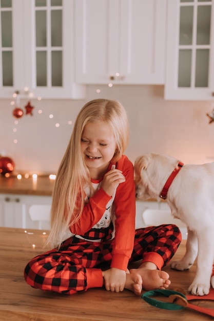 Menina loira de pijama vermelho de Natal está sentada em uma cozinha com guirlandas com um cachorro branco. fins de semana e feriados de inverno em um círculo familiar caloroso. criança e animal de estimação. espaço para texto. Foto de alta qualidade