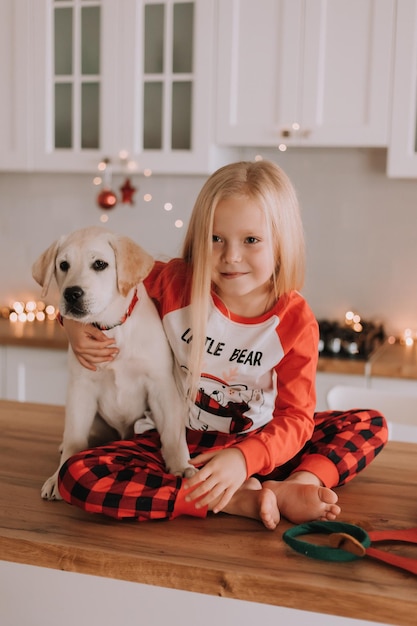 Menina loira de pijama vermelho de Natal está sentada em uma cozinha com guirlandas com um cachorro branco. fins de semana e feriados de inverno em um círculo familiar caloroso. criança e animal de estimação. espaço para texto. Foto de alta qualidade