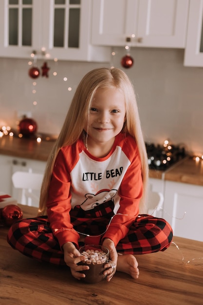 Menina loira de pijama vermelho de natal segurando uma caneca de vidro com uma bebida quente e marshmallows em uma cozinha decorada. fins de semana e feriados de inverno. estilo de vida. espaço para texto. Foto de alta qualidade