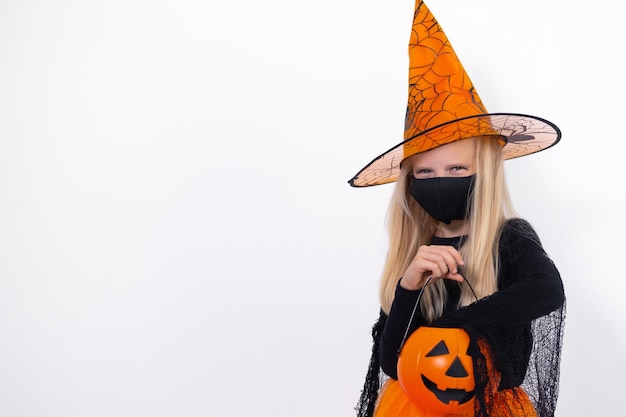 Menina loira de retrato em fantasia de bruxa usando máscara facial com abóbora de balde de doces se preparando para o Halloween no espaço de cópia de fundo branco do estúdio