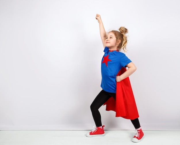 Menina loira de seis anos vestida de super-herói se divertindo em casa. Parede branca em fundo.