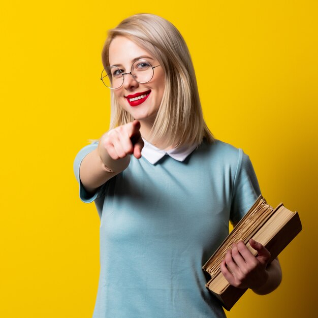 Menina loira de vestido azul e óculos com livros no espaço amarelo