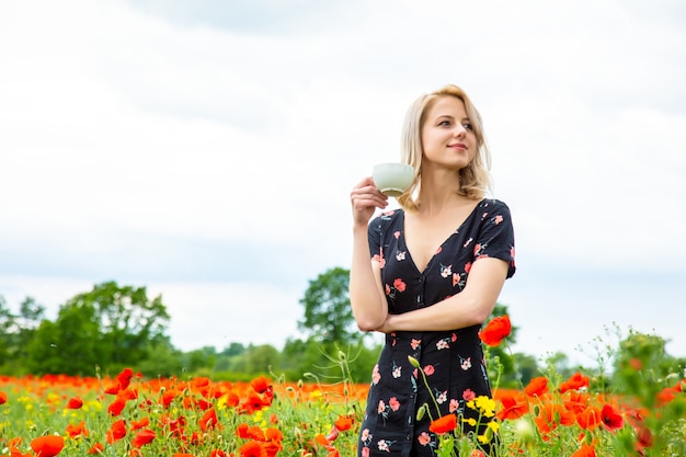 Menina loira de vestido bonito com uma xícara de café no campo de papoulas no horário de verão