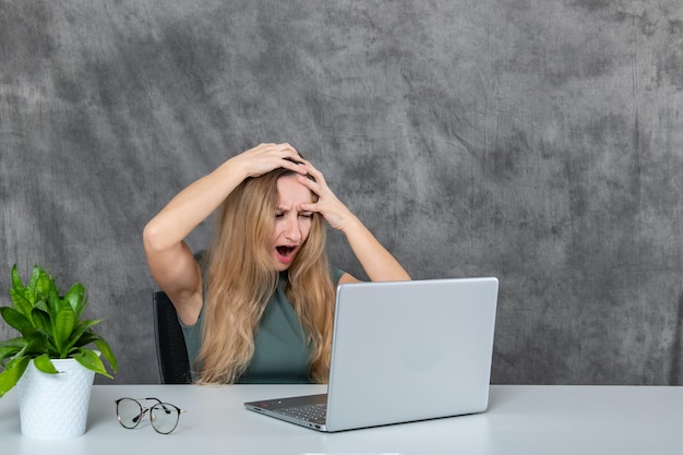 Menina loira de vestido cinza posando junto ao laptop perto de uma flor verde fresca