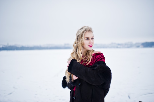 Menina loira elegância no casaco de pele e vestido vermelho posando no dia de inverno nevado.