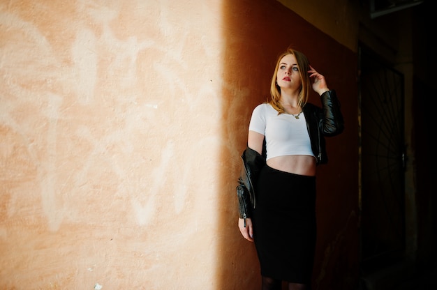 Menina loira elegante desgaste na jaqueta de couro preta posando nas ruas da cidade, velho muro com sombras.