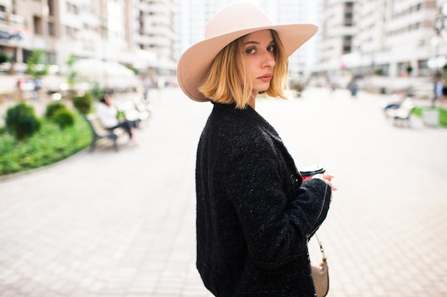 Menina loira elegante elegante de cabelo curto e chapéu posando com café sobre o fundo da rua