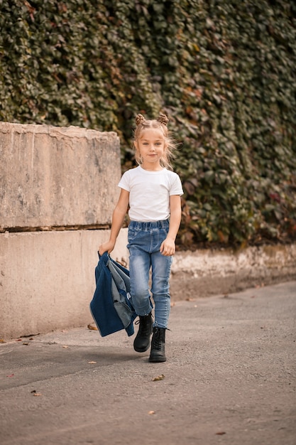 Menina loira elegante em jeans e uma camiseta branca caminha pela rua. menina de 7 anos pequena modelo, linda criança