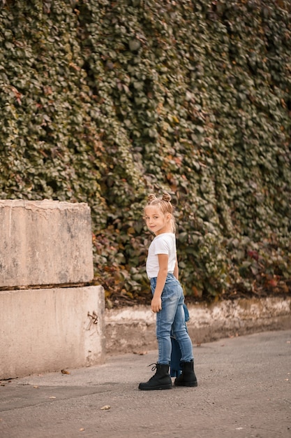 Menina loira elegante em jeans e uma camiseta branca caminha pela rua. Menina de 7 anos pequena modelo, linda criança