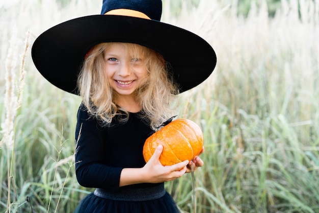 Menina loira em um terno preto e um chapéu de bruxa, o Halloween com uma abóbora nas mãos.