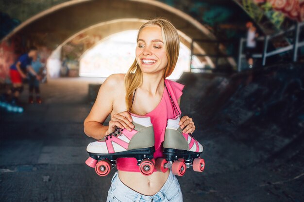 Menina loira está sorrindo. Ela está de pé em uma sala de treinamento para patins. Rolos estão pendurados no pescoço dela. Ela está segurando-os com as mãos. Menina está sorrindo e olhando para a esquerda.