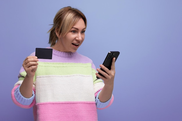 Menina loira faz uma compra na internet segurando um smartphone e um cartão de crédito com uma maquete em