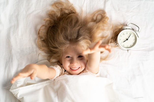 Menina loira feliz em uma cama branca com um despertador, bom dia.