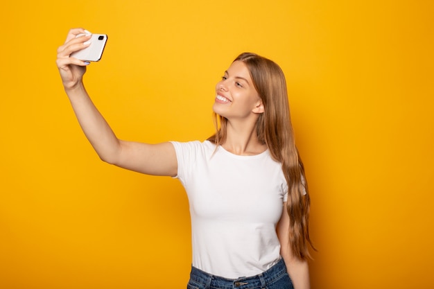 menina loira feliz tomando selfie em smartphone isolado em amarelo