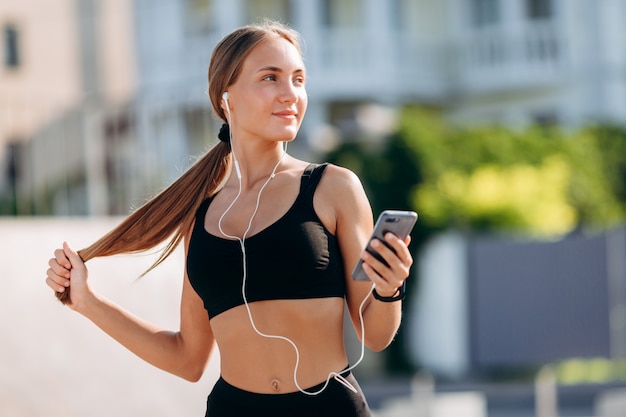 Menina loira feliz vestindo roupas esportivas em fones de ouvido segurando um smartphone.