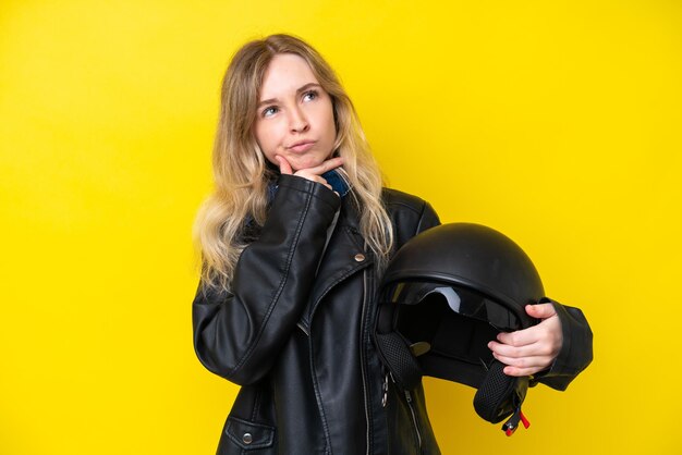 Menina loira inglesa com um capacete de motocicleta isolado em fundo amarelo com dúvidas
