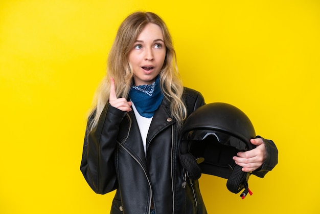 Menina loira inglesa com um capacete de motocicleta isolado em fundo amarelo pensando uma ideia apontando o dedo para cima