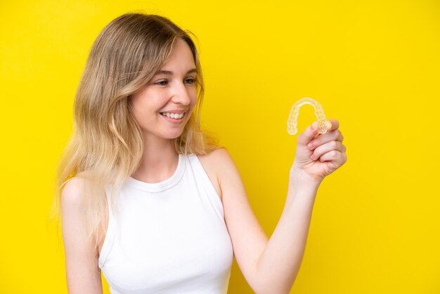 Menina loira inglesa segurando aparelho invisível isolado em fundo amarelo com expressão feliz