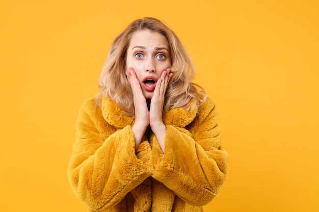 Menina loira jovem chocada com casaco de pele amarelo posando isolado no retrato de estúdio de fundo de parede laranja. Conceito de estilo de vida de emoções sinceras de pessoas. Mock up espaço de cópia. Colocando as mãos nas bochechas.