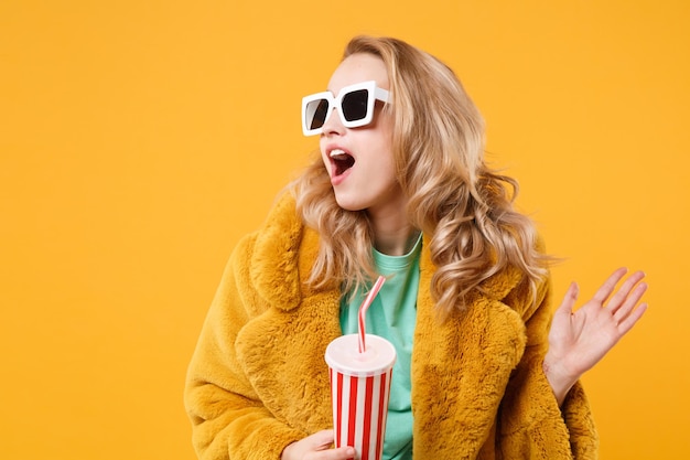 Menina loira jovem espantada com casaco de pele amarelo, óculos escuros posando isolado em fundo laranja. Conceito de estilo de vida de pessoas. Mock up espaço de cópia. Segure o copo plástico de cola ou refrigerante espalhando as mãos.