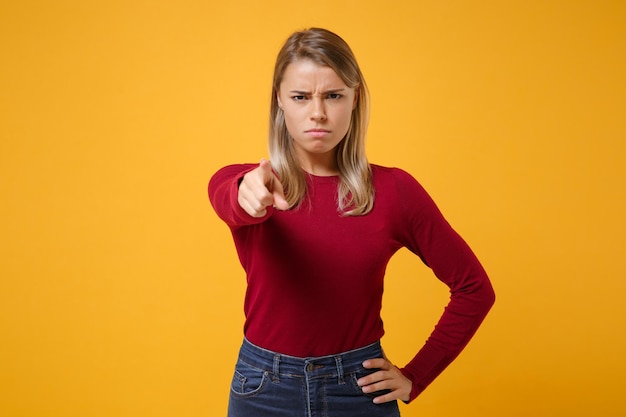 Menina loira jovem estrita insatisfeita em roupas casuais posando isolada no retrato de estúdio de fundo amarelo laranja. Conceito de estilo de vida de pessoas. Simule o espaço da cópia. Aponte o dedo indicador para a câmera.