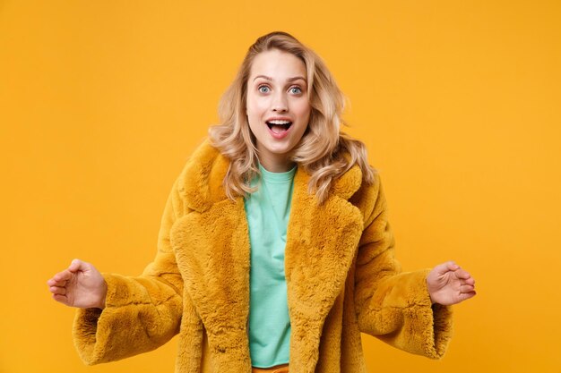 Menina loira jovem excitada com casaco de pele amarelo posando isolado em fundo laranja no estúdio. Conceito de estilo de vida de pessoas. Mock up espaço de cópia. Gesticulando demonstrando tamanho com espaço de trabalho horizontal.