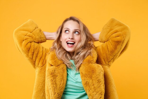 Menina loira jovem pensativa com casaco de pele amarelo posando isolado em fundo laranja, retrato de estúdio. Conceito de estilo de vida de emoções de pessoas. Mock up espaço de cópia. De mãos dadas atrás da cabeça, olhando para cima.