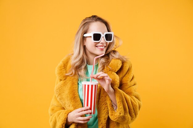 Menina loira jovem sorridente com casaco de pele amarelo, óculos escuros posando isolado em fundo laranja no estúdio. Conceito de estilo de vida de pessoas. Mock up espaço de cópia. Segurando o copo plástico de cola ou refrigerante.