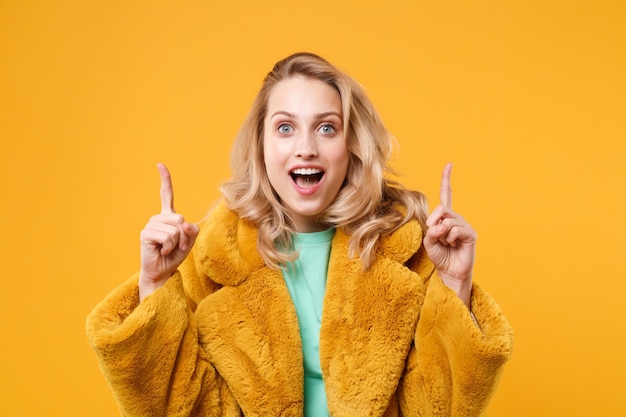 Menina loira jovem surpresa com casaco de pele amarelo posando isolado no retrato de estúdio de fundo de parede laranja. Conceito de estilo de vida de emoções sinceras de pessoas. Mock up espaço de cópia. Aponte os dedos indicadores para cima.