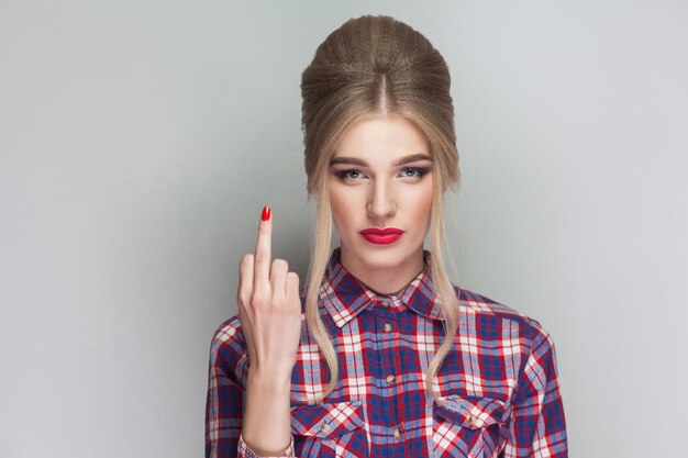 Menina loira linda com raiva de camisa quadriculada rosa e penteado updo coletado em pé, olhando para a câmera e mostrando o dedo médio com cara de pôquer. tiro de estúdio interno. isolado em fundo cinza