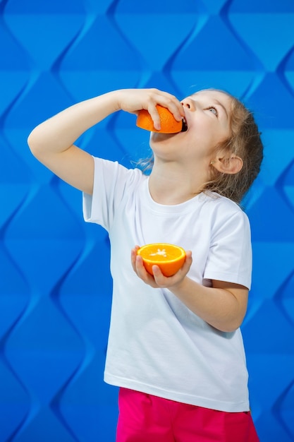 Menina loira muito bonita em uma camiseta branca sorri sobre um fundo azul. criança bonita elegante tem meia laranja nas mãos. o conceito de um retrato emocional.