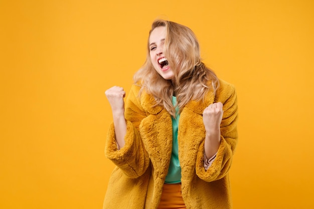 Menina loira muito feliz com casaco de pele amarelo posando isolado no retrato de estúdio de fundo de parede laranja. Conceito de estilo de vida de emoções sinceras de pessoas. Mock up espaço de cópia. Fazendo o gesto do vencedor.