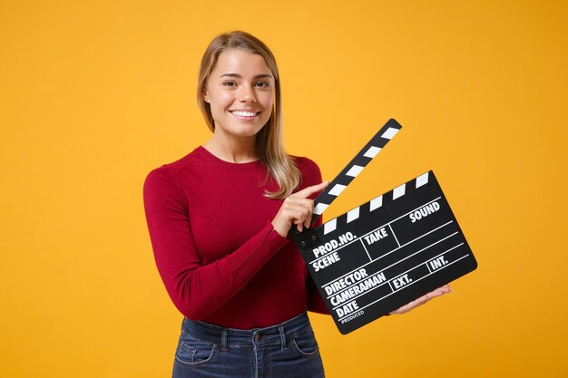 Menina loira muito jovem em roupas casuais posando isolada no fundo da parede laranja amarela no estúdio. Conceito de estilo de vida de pessoas. Simule o espaço da cópia. Segure a claquete de filmagem preta clássica.