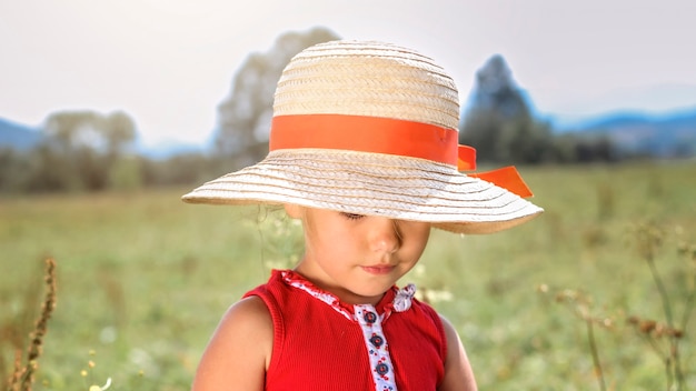 Menina loira pré-escolar em um chapéu de palha e vestido vermelho no prado em dia de sol de verão