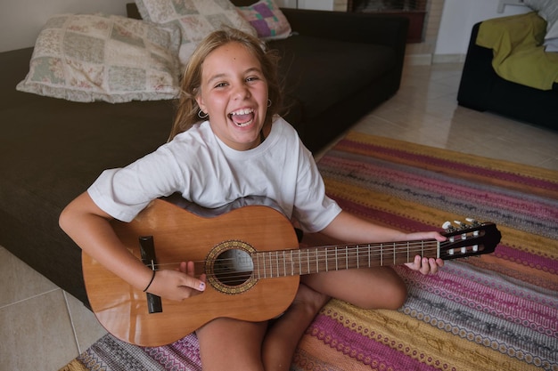 Menina loira sentada no chão de uma casa tocando guitarra enquanto está cantando e olhando para a câmera